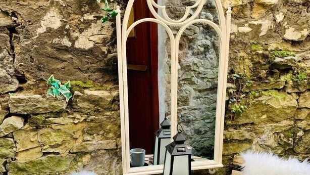 Un mur en pierre, une table en bois et deux chaises blanches à l’assise moelleuse. Un miroir avec un cadre en bois est fixé au mur.