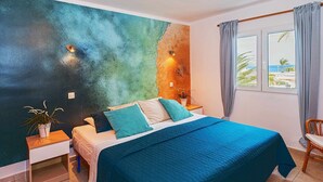Een slaapkamer met een bed, nachtkastjes, een kamerplant en uitzicht op zee door een raam.
