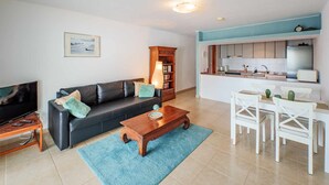 Een moderne woonkamer met een zwarte bank, een houten salontafel en een televisie op een meubel. Er is een keukenhoek met witte kasten en een blauw aanrecht.