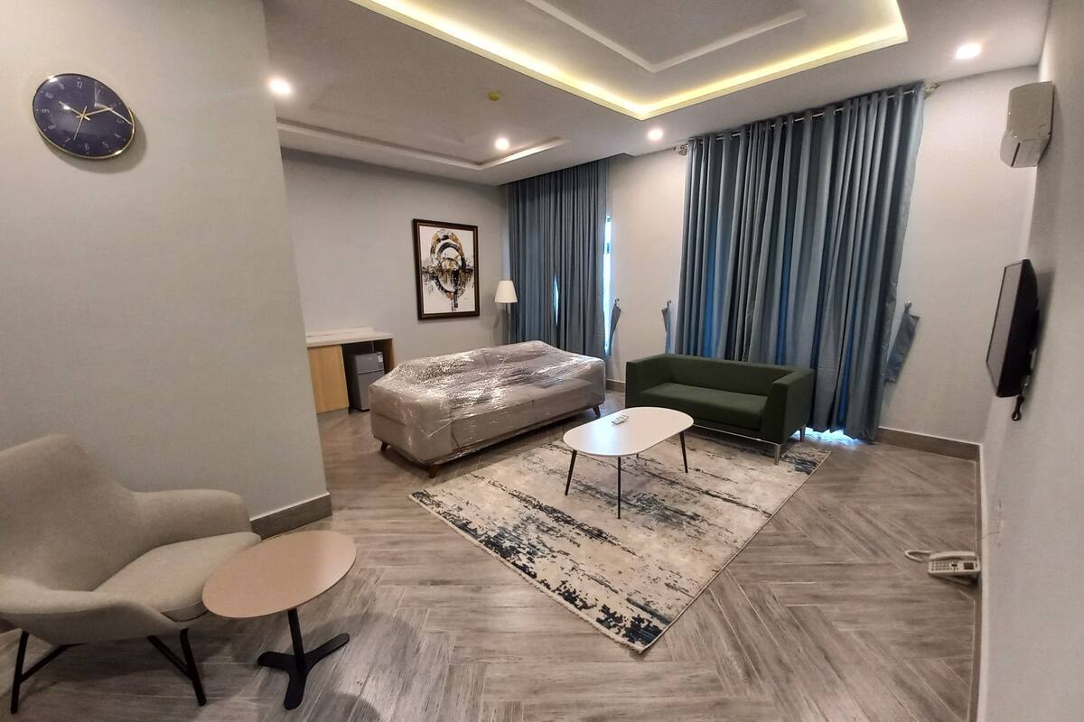 A modern living room with a sofa, a small table, a chair, a bed, a clock, and a TV.