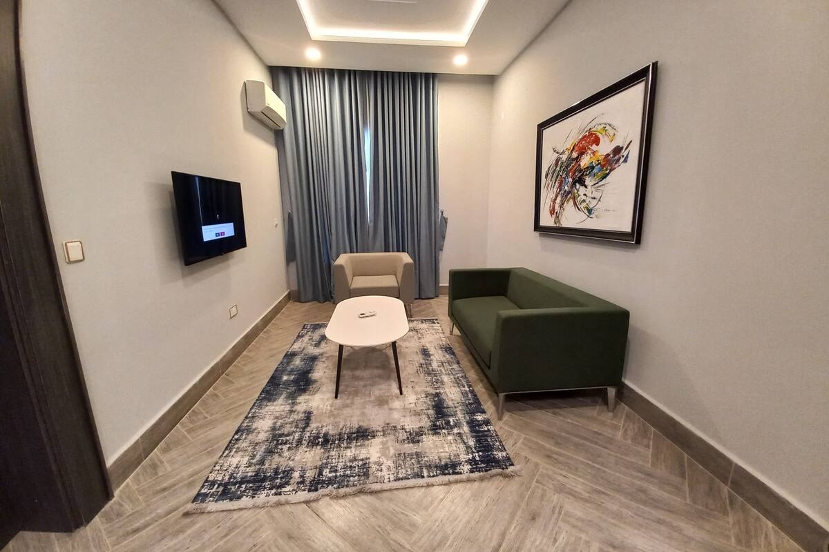 A modern living room with a flat-screen TV, a green sofa, a beige armchair, and a patterned rug.