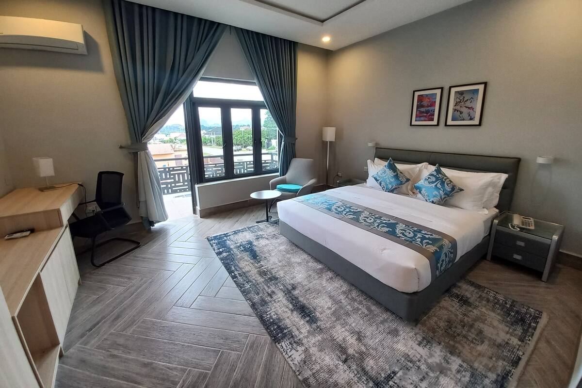 A hotel room with a bed, a desk, a chair, a small table, a nightstand, and a window with curtains.