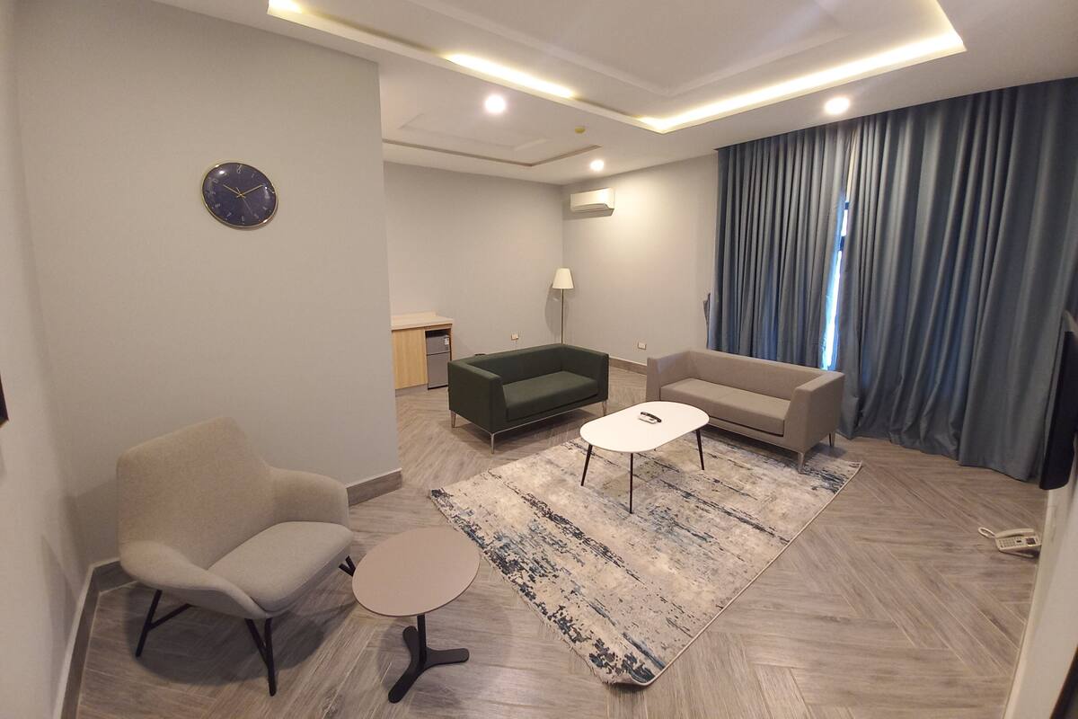 A modern living room with a sofa, armchair, coffee table, and a small kitchen area in the background.