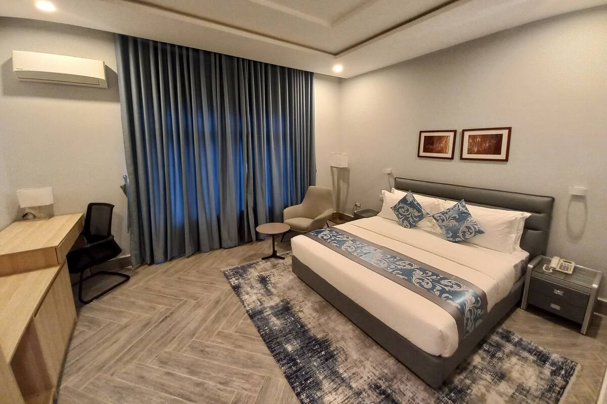 A modern hotel room with a large bed, a desk, a chair, and a small table.