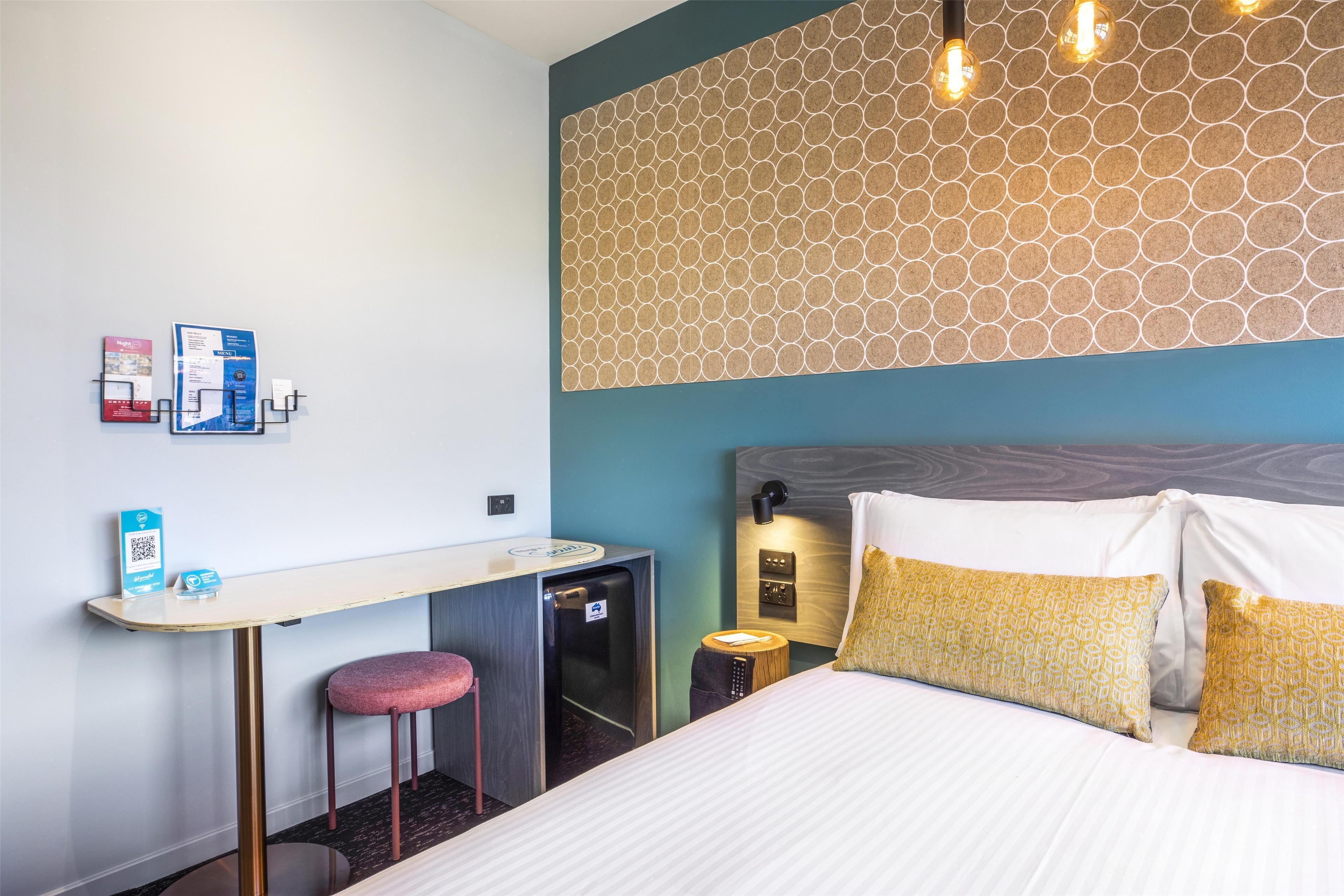 Una habitación de hotel con una cama, un escritorio, un refrigerador pequeño, un taburete y una lámpara fijada a la pared.