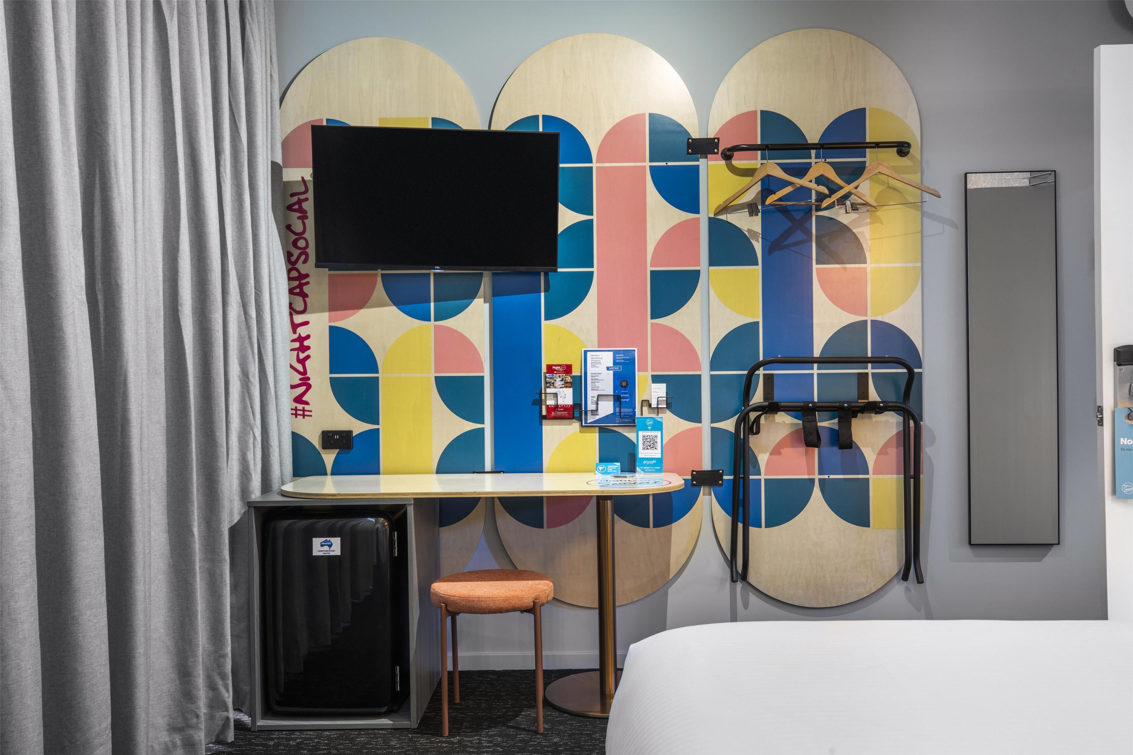 A hotel room with a bed, a desk, a chair, a TV mounted on a colorful wall, a coat rack, and a small refrigerator.