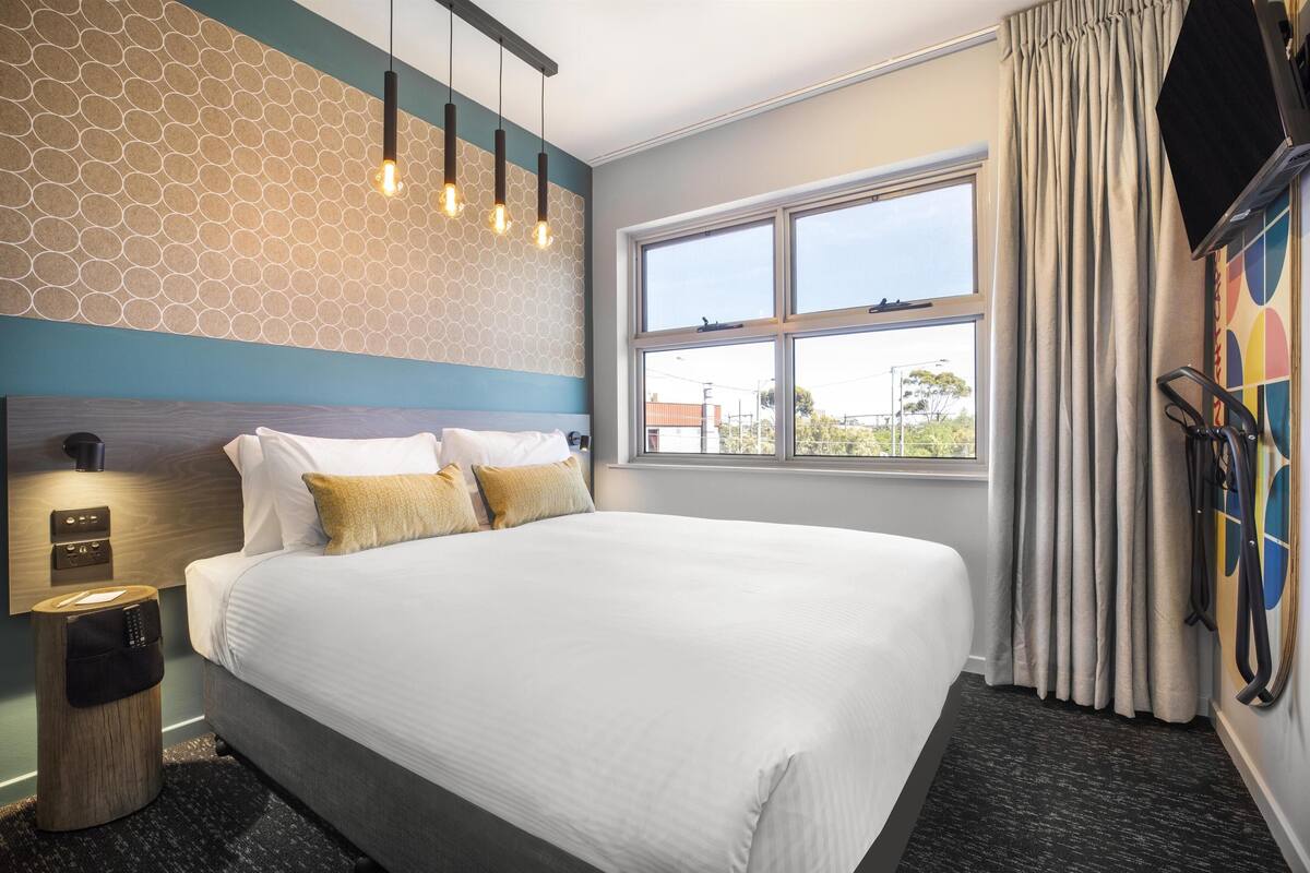 A hotel room with a large bed, a window with curtains, a wall with a geometric pattern, and a mounted television.