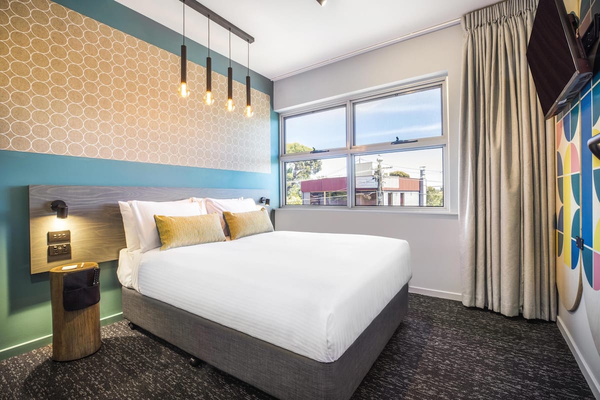 A hotel room with a large bed, a nightstand with a lamp, a wall-mounted TV, and a window with curtains.