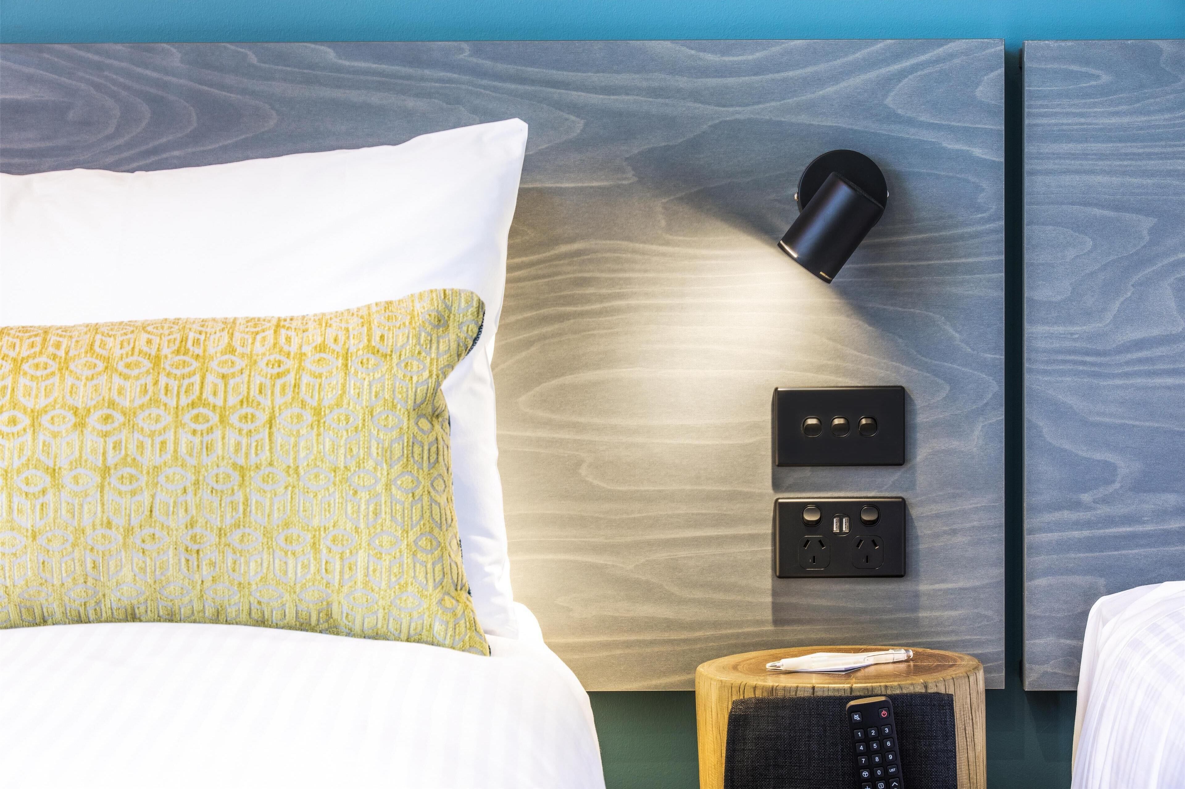 Habitación de hotel con cama, mesita de noche con control remoto, lámpara de pared y enchufe.
