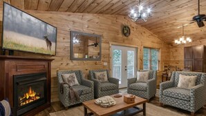 L’intérieur chaleureux d’un chalet avec une cheminée, des fauteuils à motifs, un plafond en bois et un téléviseur fixé au mur.