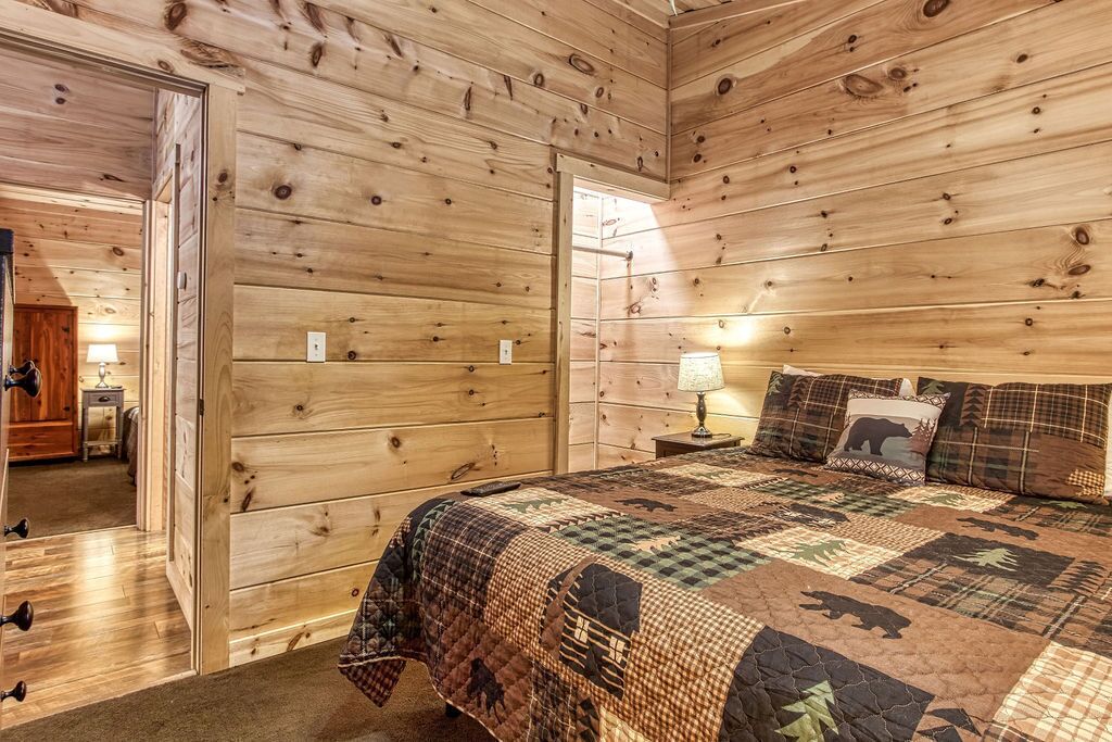 A wooden-paneled bedroom with a quilted bedspread, a nightstand with a lamp, and a door leading to another room.