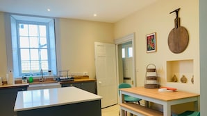 A modern kitchen with a central island, a dining area with a bench, and a view of the adjacent living space. - The Garden Flat - An Elegant Georgian Single Level Holiday Home (Charmouth)
