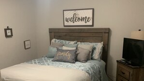 Una camera da letto con un letto, un comodino con una lampada, un cartello con la scritta 'welcome' incorniciata e un orologio appeso al muro.