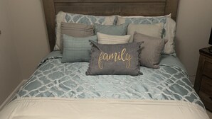 Un letto rifatto con cura, con una coperta a motivi e diversi cuscini, tra cui uno con la scritta 'family' ricamata. Sopra il letto, un cartello incorniciato con la scritta 'welcome'.