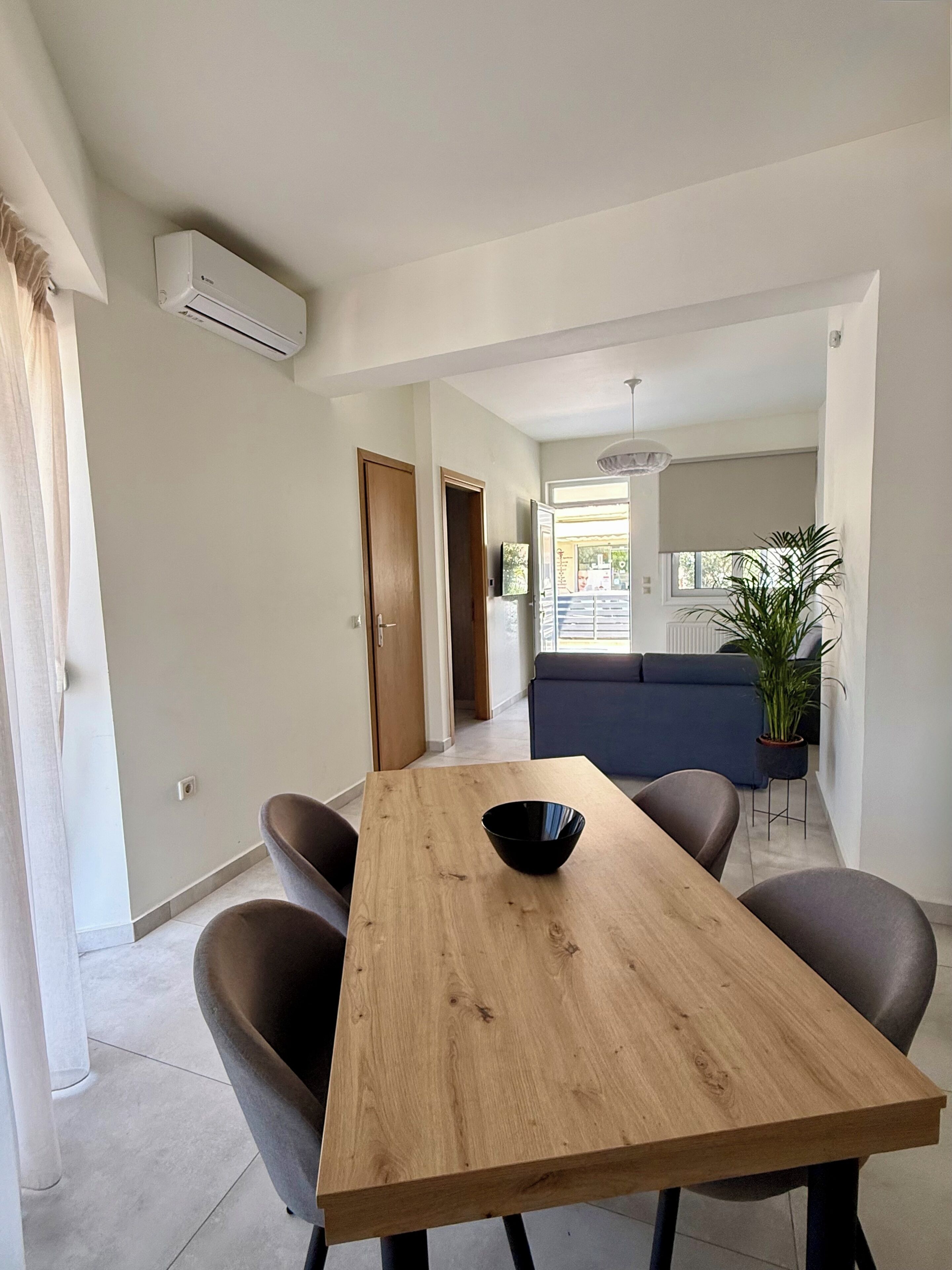 Een eetgedeelte met een houten tafel, stoelen en een schaal. Achtergrond toont een bank en een plant.