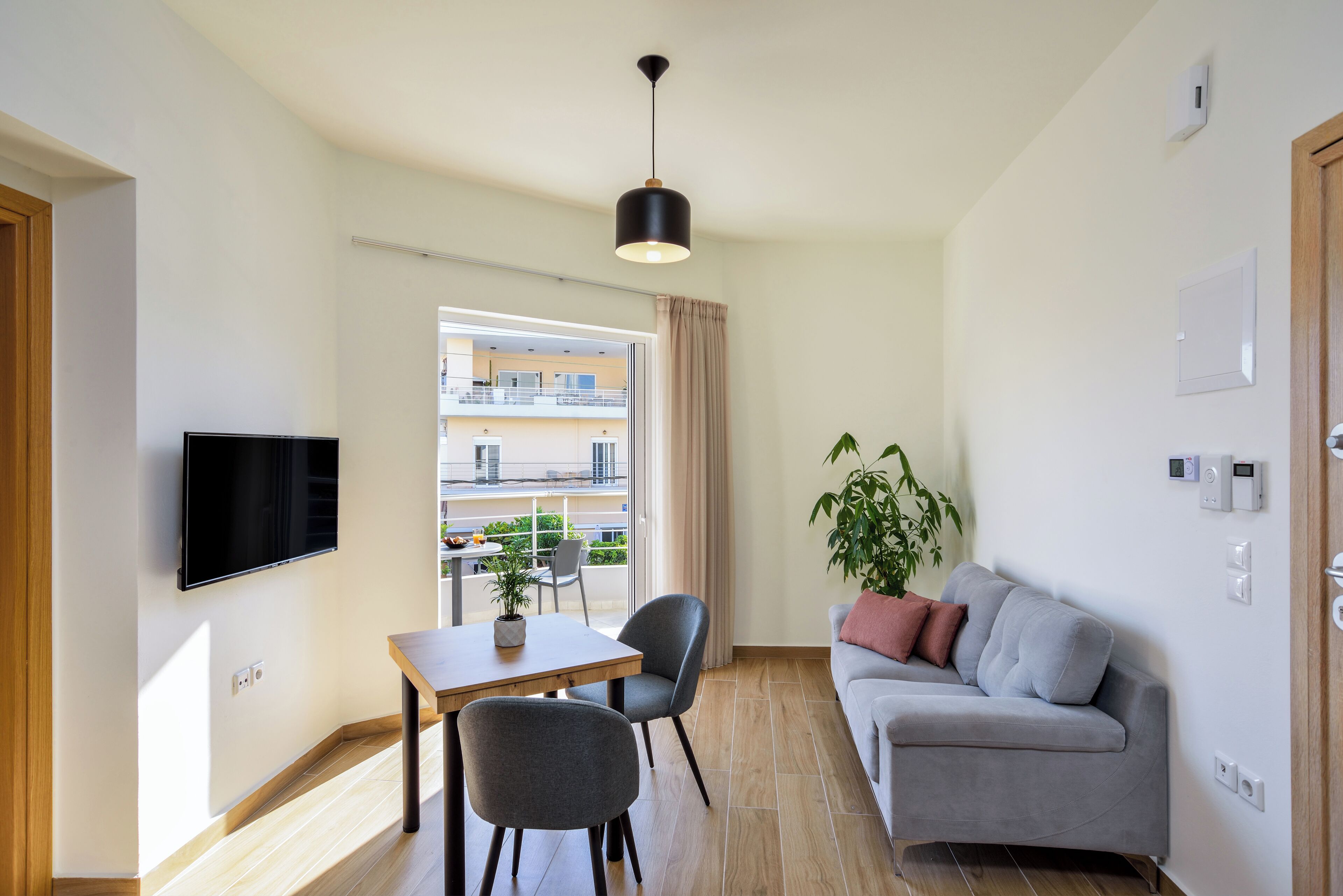 A modern living room with a flat-screen TV, a dining table with chairs, a grey sofa, a potted plant, and a balcony with outdoor furniture.