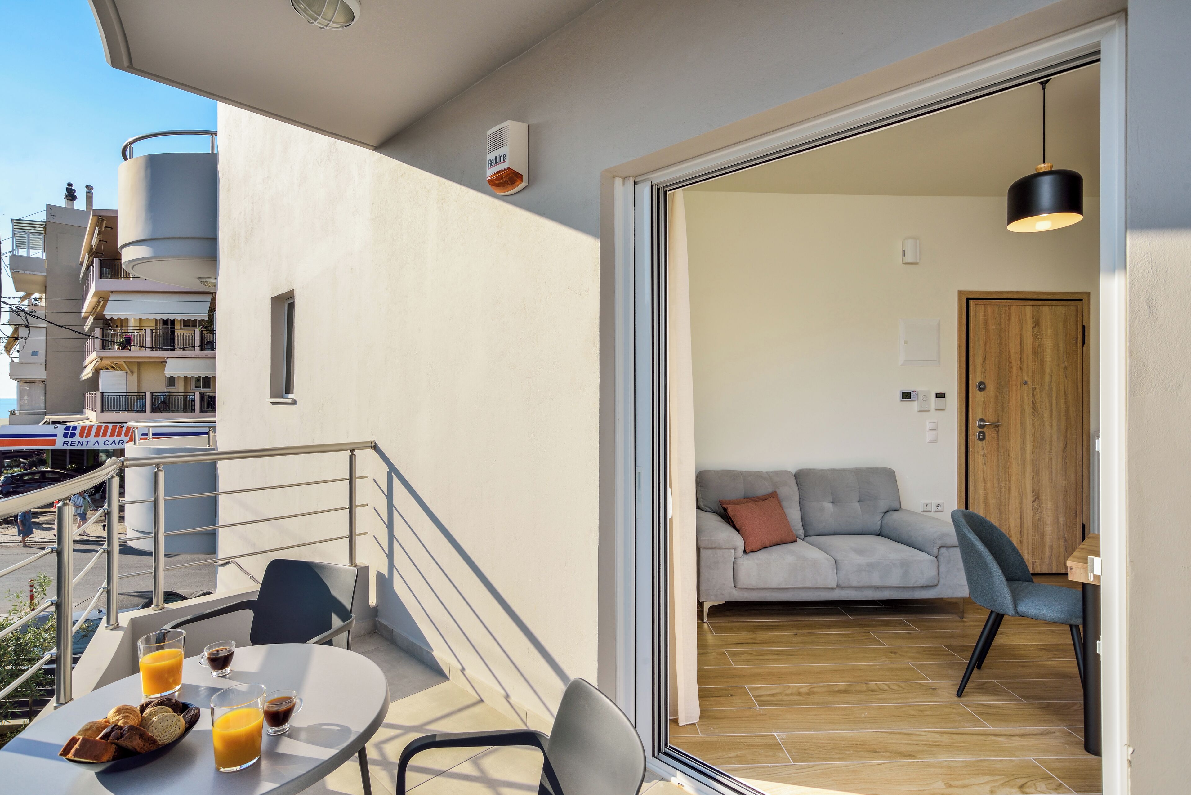 Un balcón con mesa, sillas y alimentos para el desayuno; una sala de estar con sofá y puerta; y la fachada de un edificio con un cartel de 'Rent a Car'.