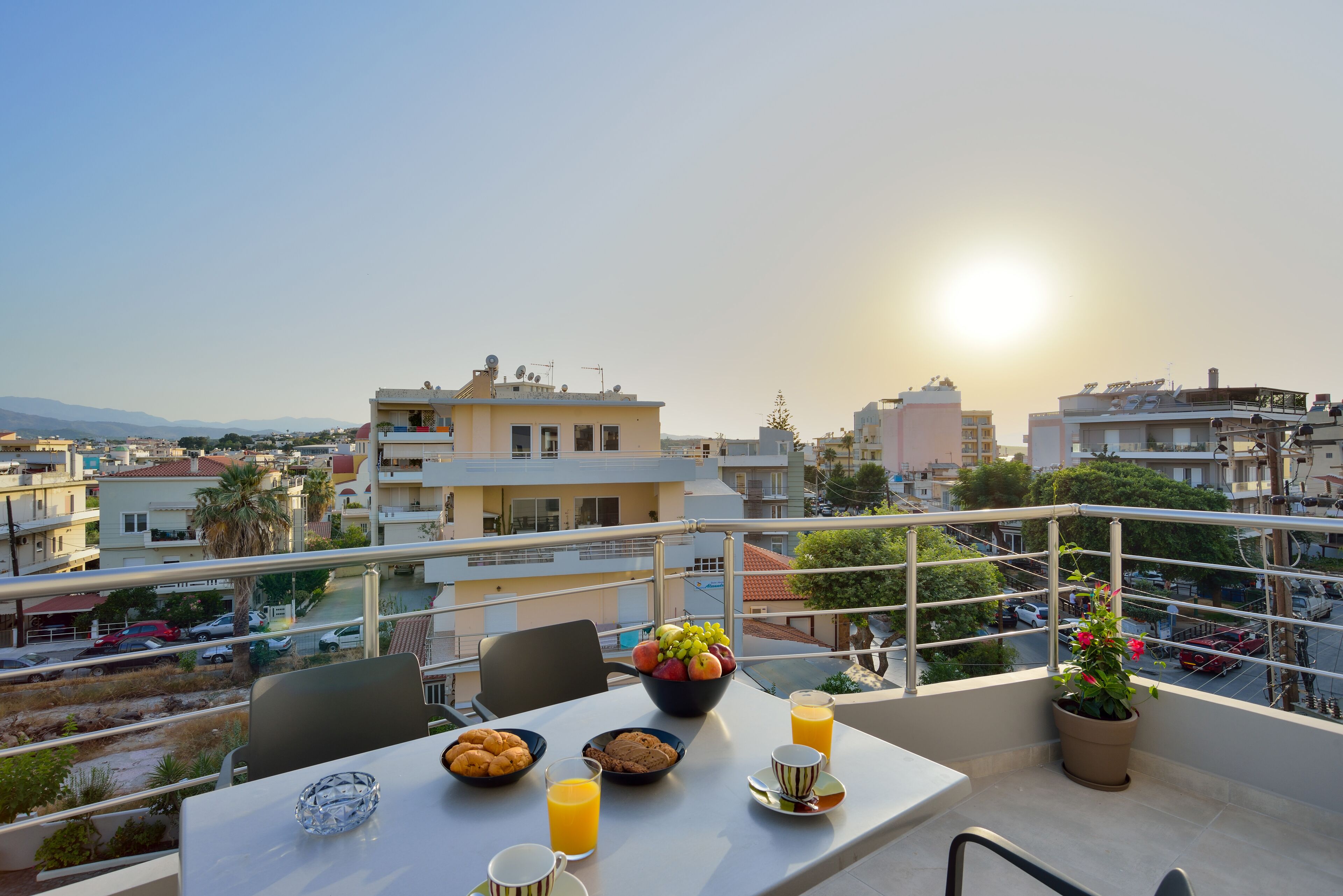Un balcón con una mesa lista para el desayuno, que incluye frutas, galletas y jugo, con vista a un paisaje urbano con edificios y cielo despejado.