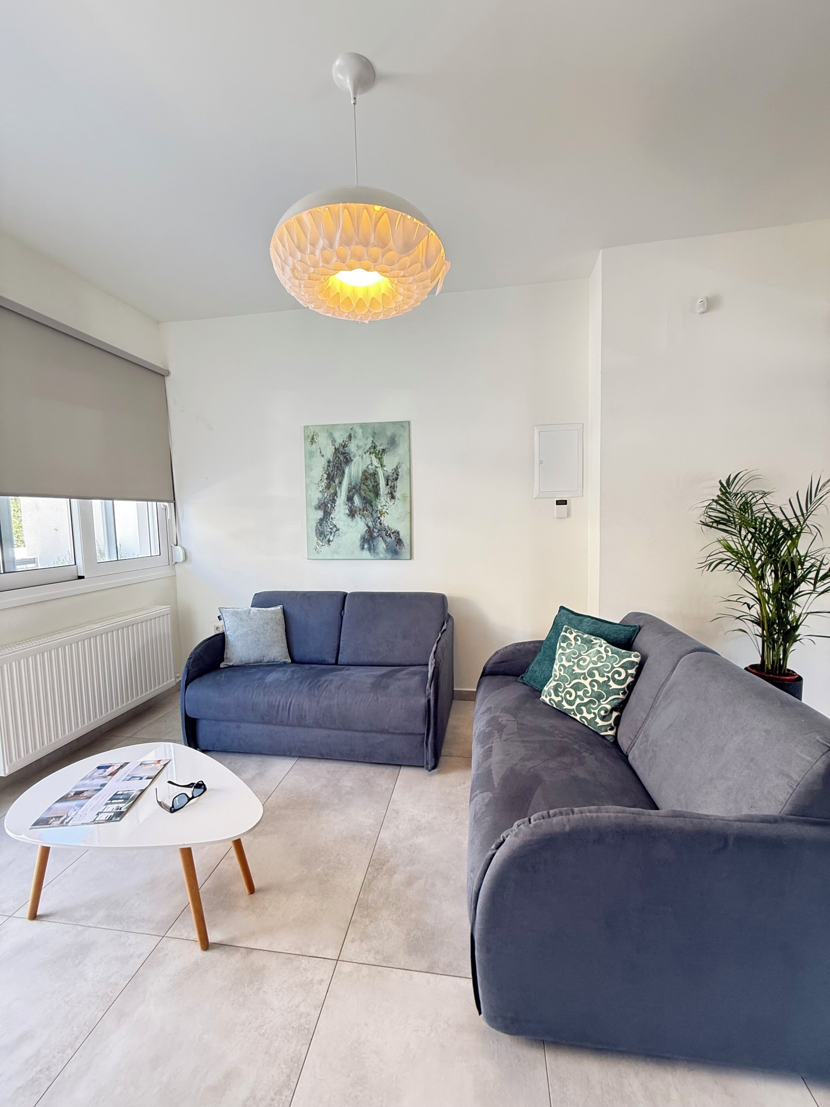 A modern living room with two grey sofas, a white coffee table, a hanging light fixture, a wall-mounted abstract painting, and a potted plant.