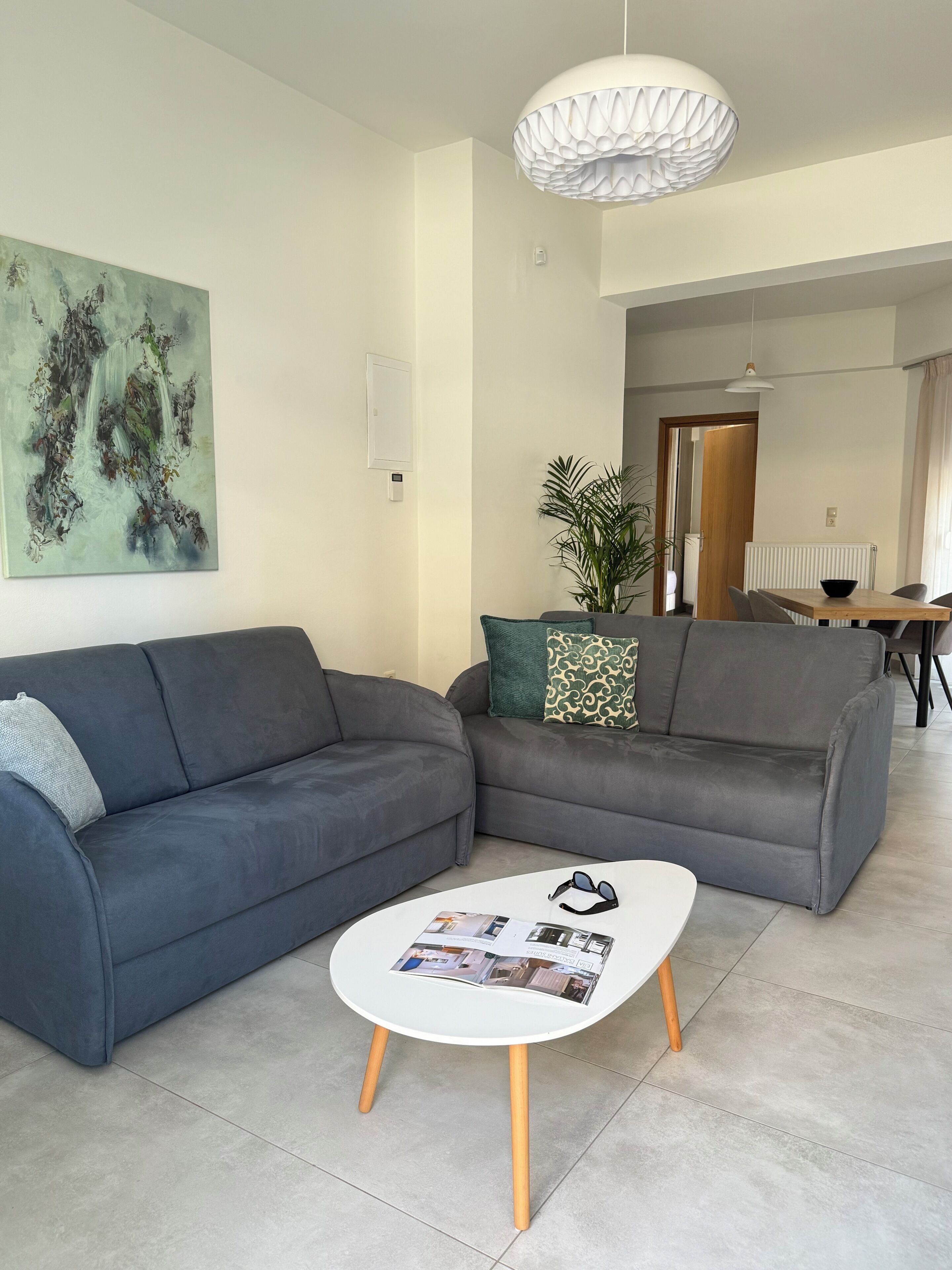 A modern living room with a grey sectional sofa, a white coffee table with magazines, a large abstract painting, and a hanging light fixture.