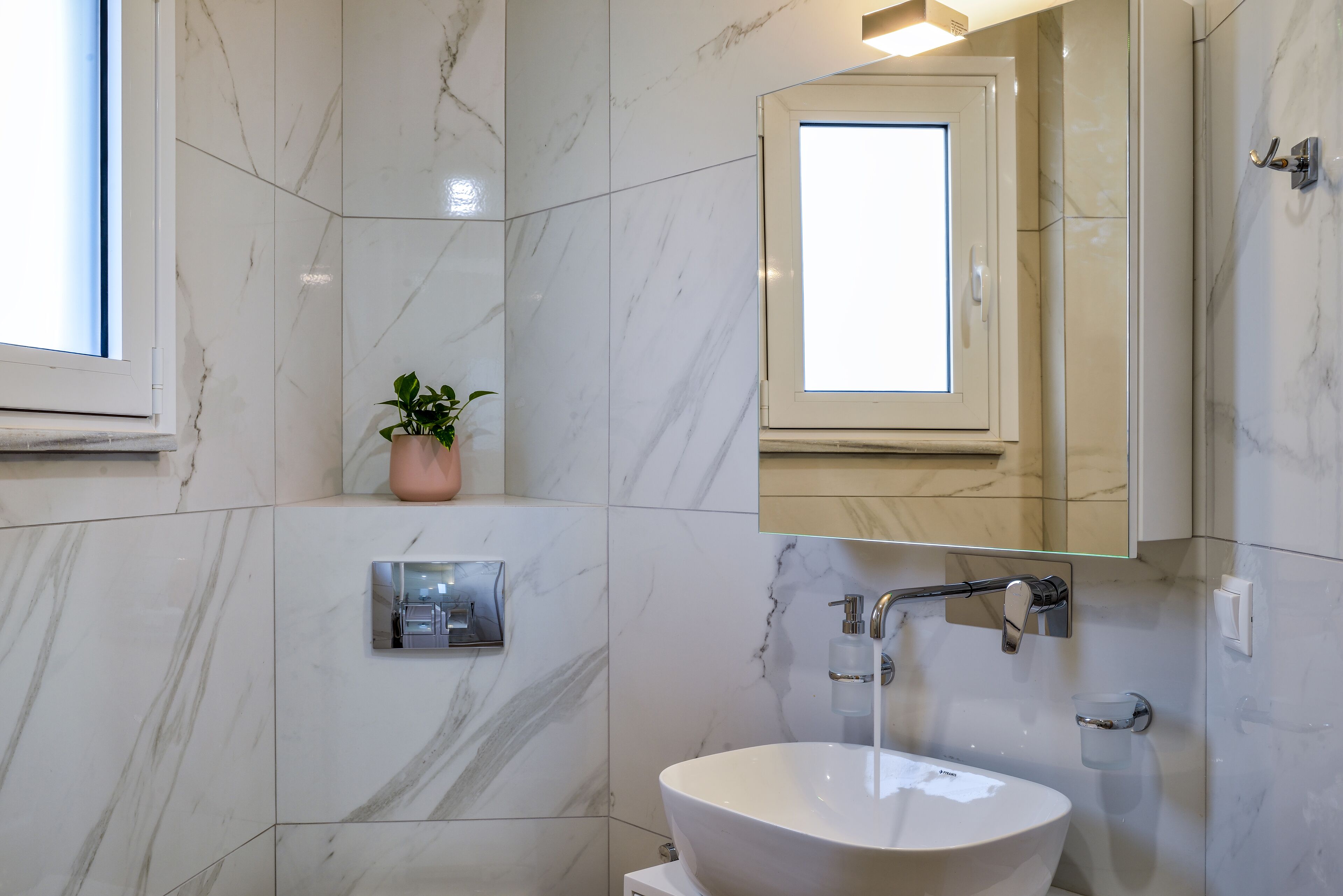 Une salle de bain avec un lavabo en marbre blanc, une plante en pot, un sèche-mains et un miroir.