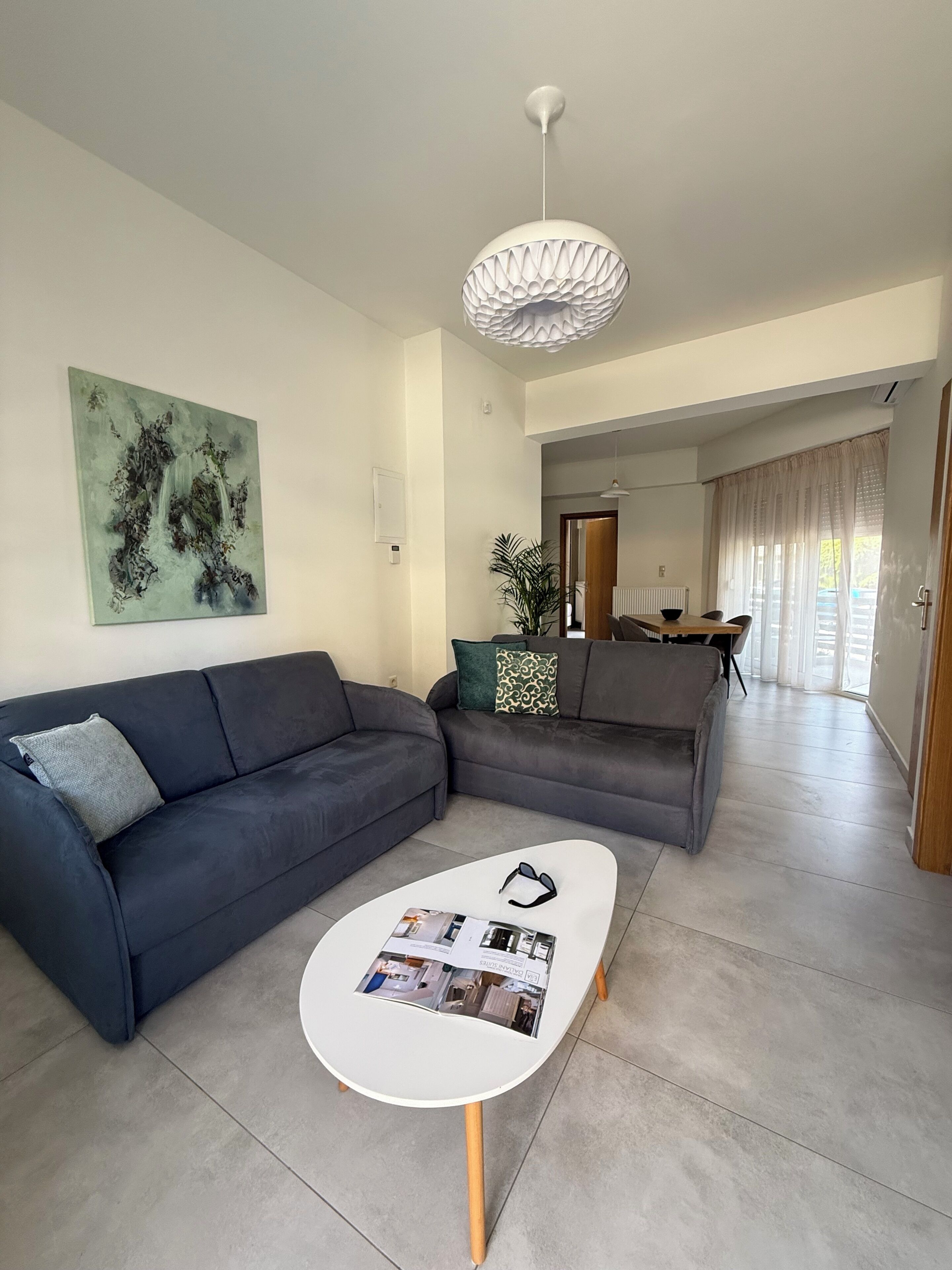 A modern living room with a dark grey sofa, a white coffee table with magazines, a green abstract painting, and a ceiling light fixture.
