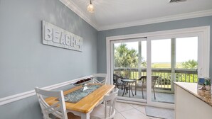 Een eetruimte met een tafel en stoelen, een 'BEACH' bord op de muur, en uitzicht op palmbomen door grote glazen deuren.