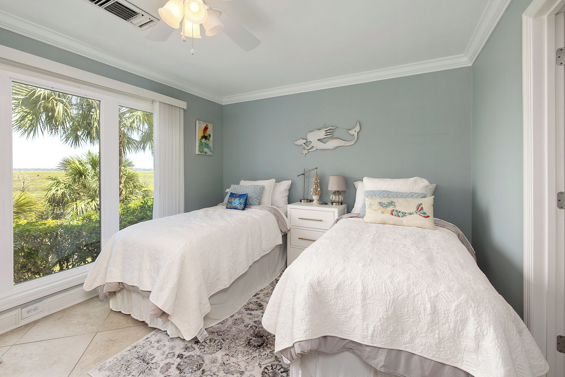 A bedroom with two beds, a ceiling fan, a nightstand with a lamp, and a view of greenery outside.