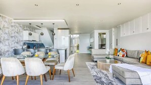 A modern living room with a grey sofa, a dining area with a glass table, and a kitchen in the background.