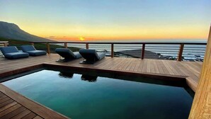 Une terrasse en bois avec une piscine et des chaises longues, offrant une vue sur l’océan au coucher du soleil.