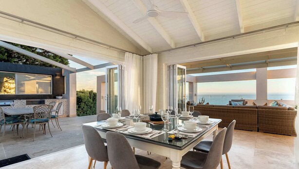 Un espace repas moderne avec une table en verre, des chaises grises et une vue sur la mer.
