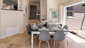 On aperçoit un coin repas avec une table en verre dressée pour six personnes, entourée de chaises grises. Au fond, on distingue une cuisine, et un escalier mène à un étage supérieur.