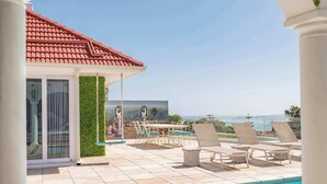 A pool area with a clear view of the sea, lounge chairs, and a building with a red roof.