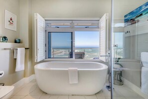 A modern bathroom with a freestanding bathtub, a glass shower enclosure, and a view of the ocean through a large window.