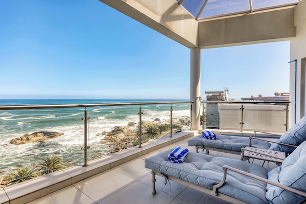 A balcony with a view of the ocean, a seating area with cushions, and a glass railing.