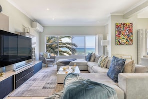 A modern living room with a large flat-screen TV, a sofa, a coffee table, and a view of the ocean.