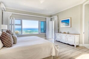 A bedroom with a large bed, a view of the ocean, a white dresser, and a framed picture on the wall.