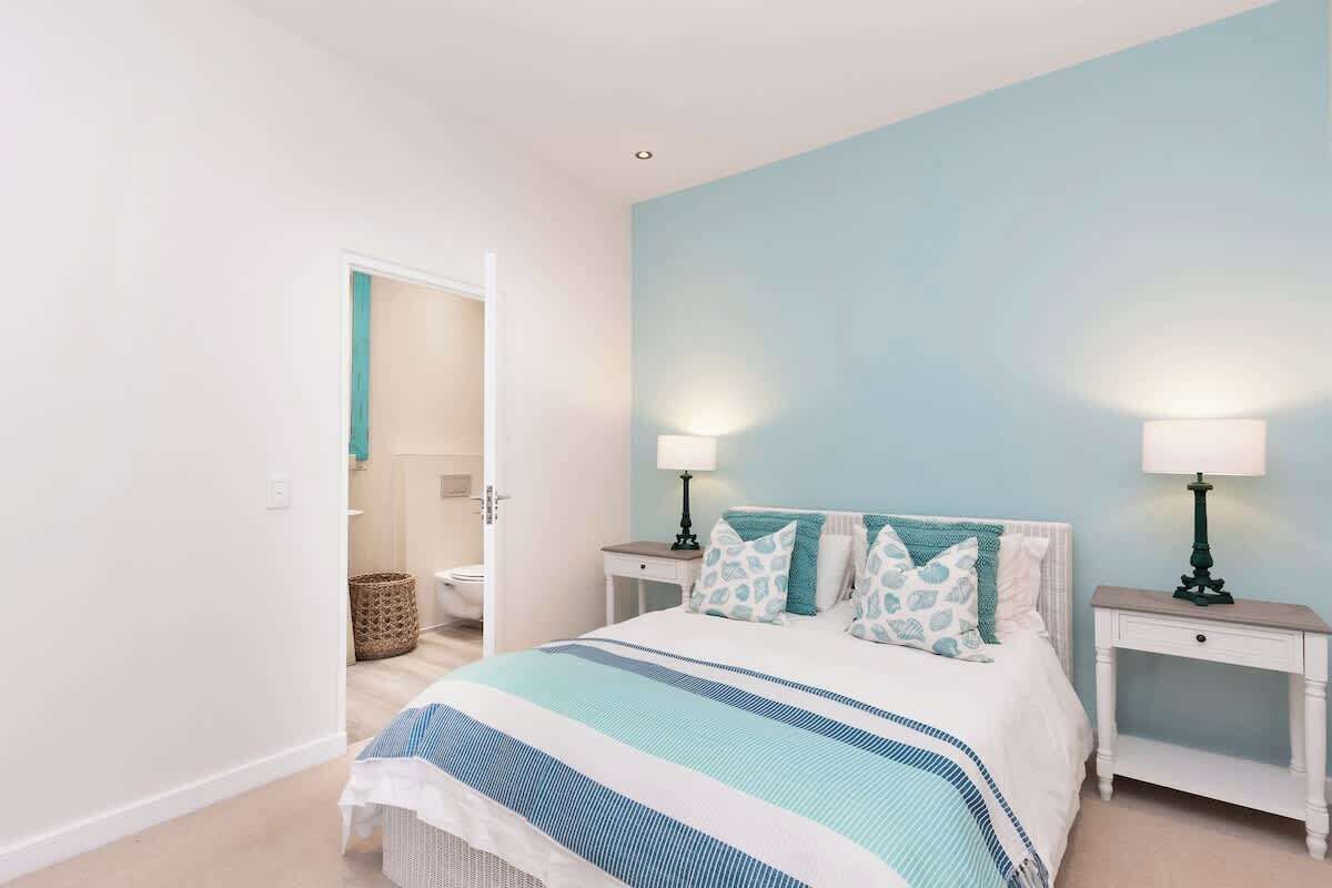A bedroom with a bed, two bedside tables with lamps, a bathroom visible through an open door, and a light blue wall.