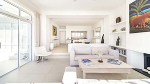 Un salon moderne avec un canapé blanc, une table basse sur laquelle sont posés des livres, et un coin repas à l’arrière-plan.