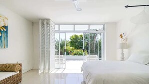 Une chambre à coucher lumineuse et moderne, dotée d’un grand lit, d’un canapé blanc et offrant une vue sur un jardin grâce à de grandes portes vitrées.