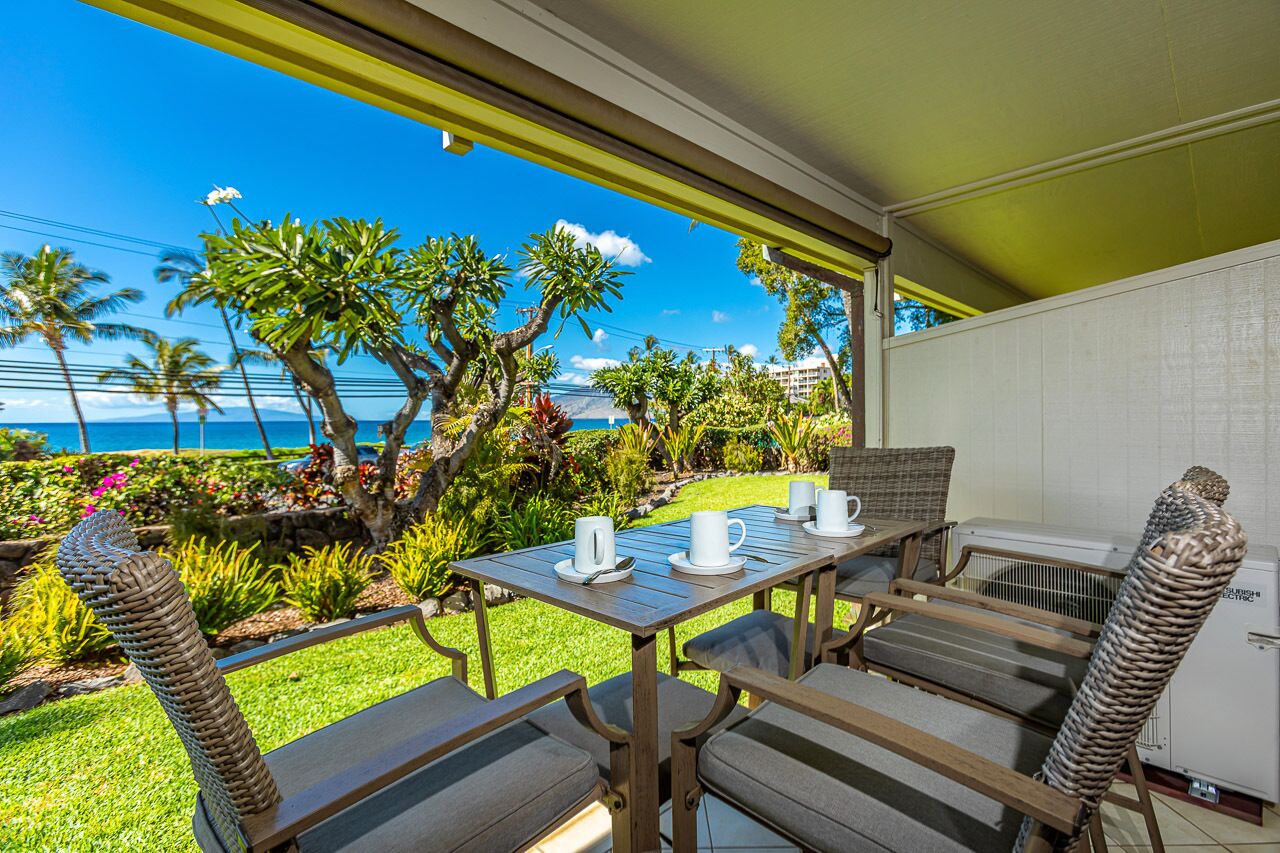 A covered outdoor dining area with a table and chairs, overlooking a garden with palm trees and a view of the ocean.