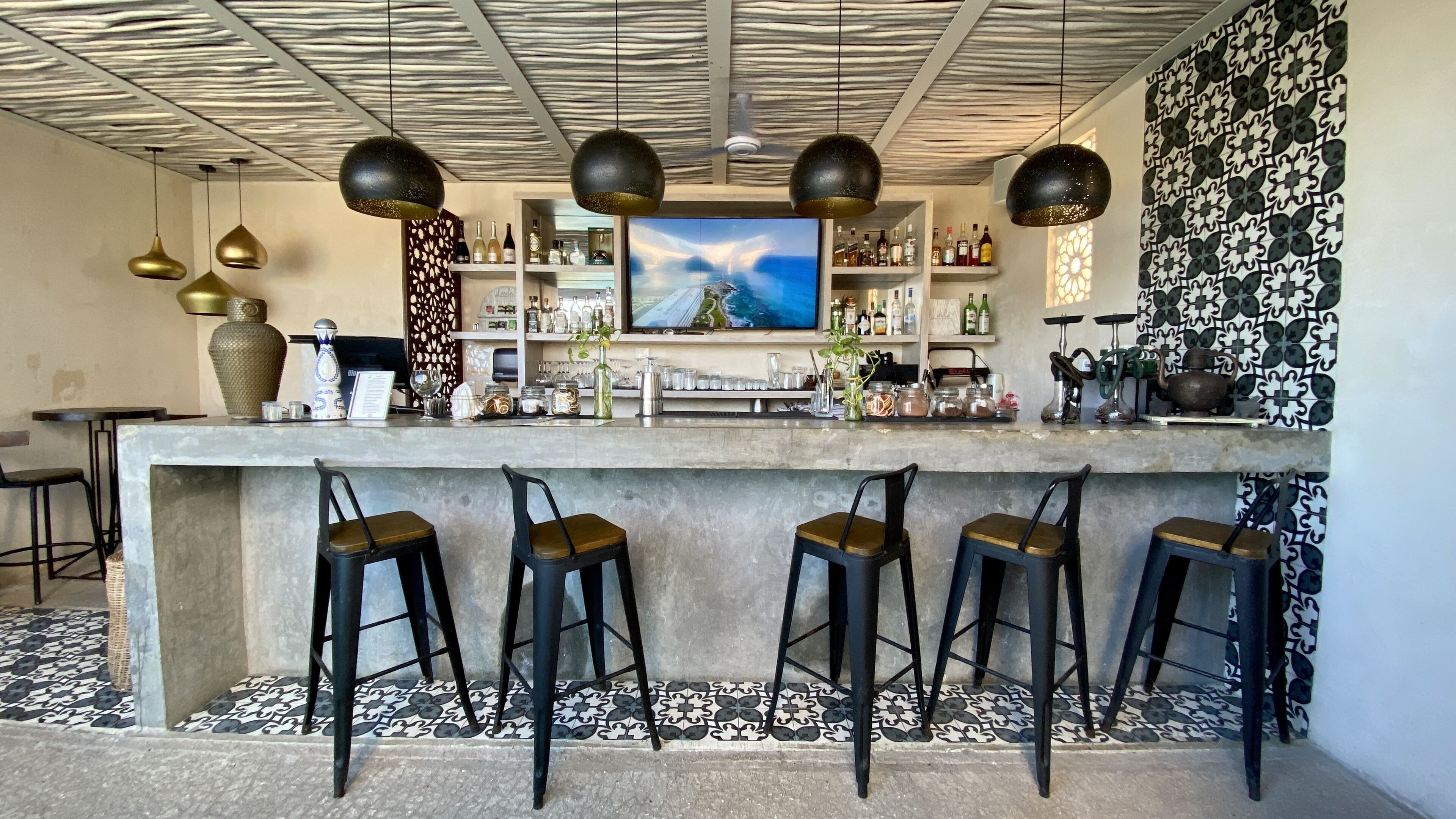 A bar area with a concrete counter, black bar stools, and a large TV screen displaying a scenic view.