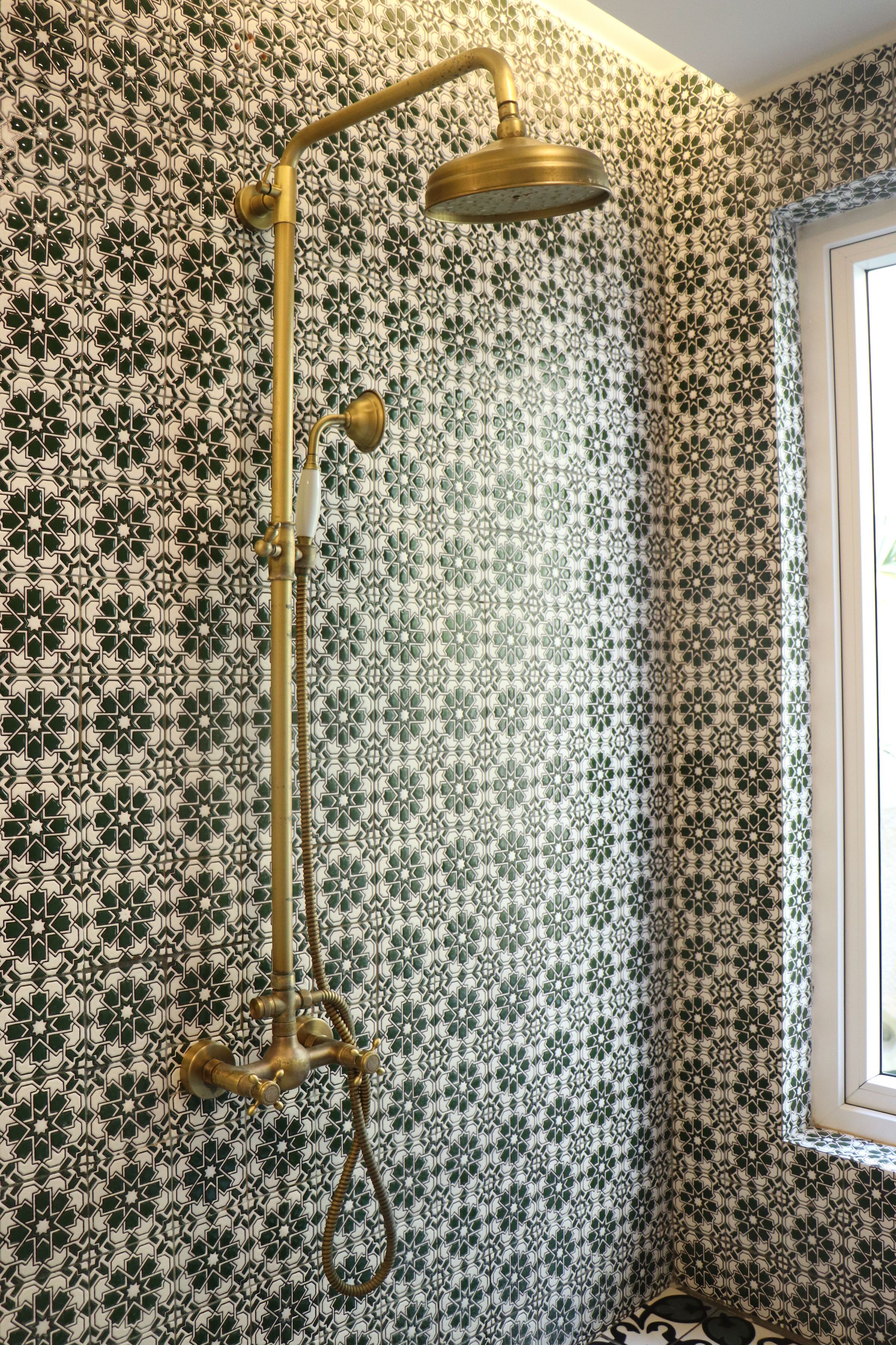 A bathroom with a patterned wall, a shower with a gold fixture, and a window with daylight.