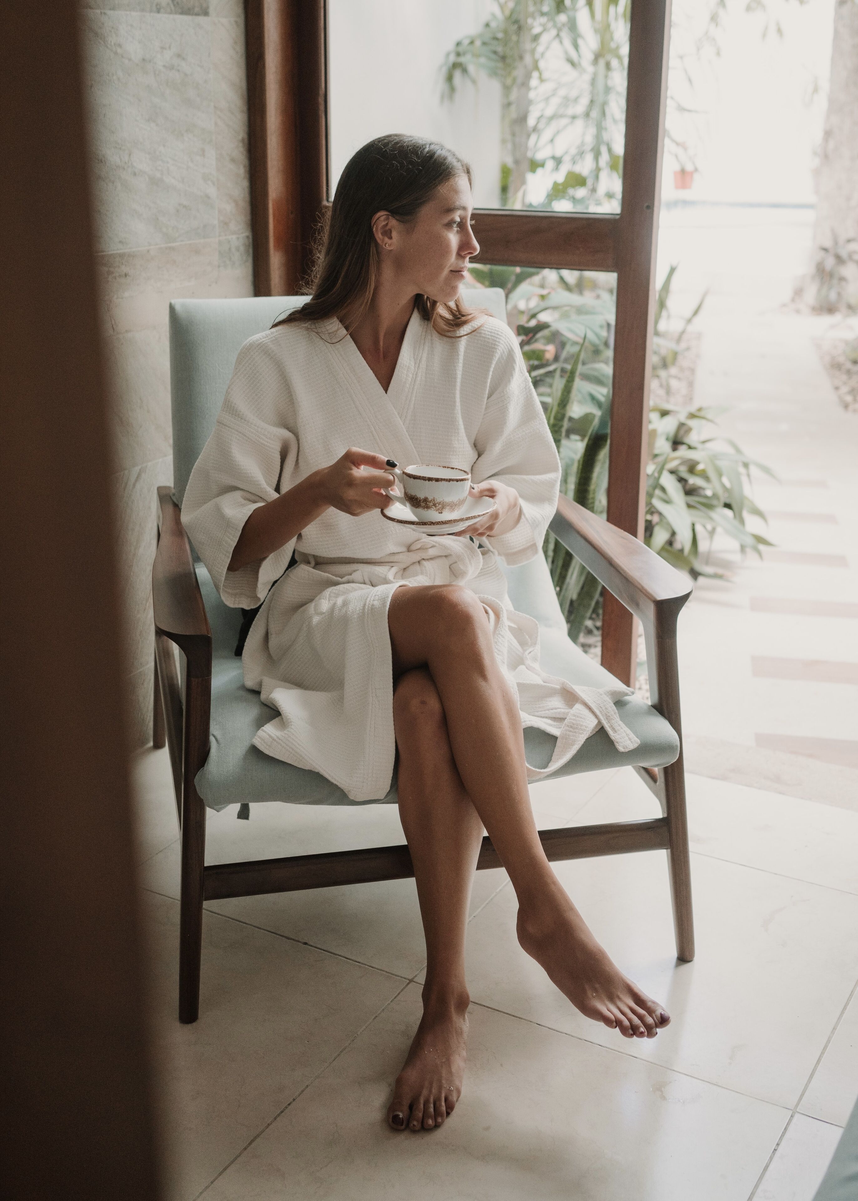 A person sitting on a wooden chair in a room with a tiled floor and a window with a view of plants outside.