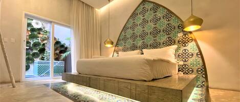 A modern bedroom with a large bed, patterned headboard, and a view of a balcony with plants.