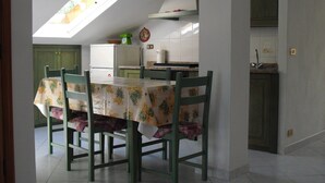 A dining area with a table covered by a floral tablecloth, green chairs, a kitchen in the background, and a skylight above.