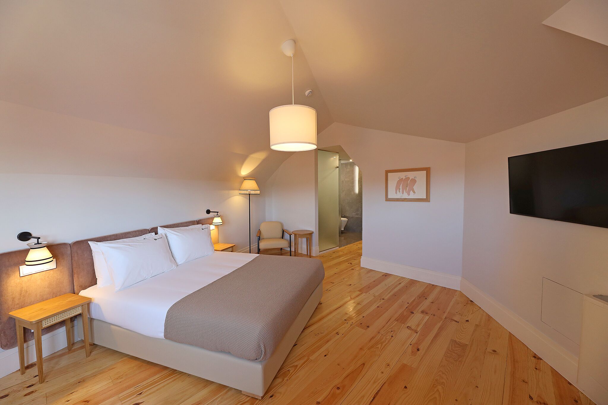 A bedroom with a bed, bedside lamps, a small table, a chair, a TV, and a wooden floor.