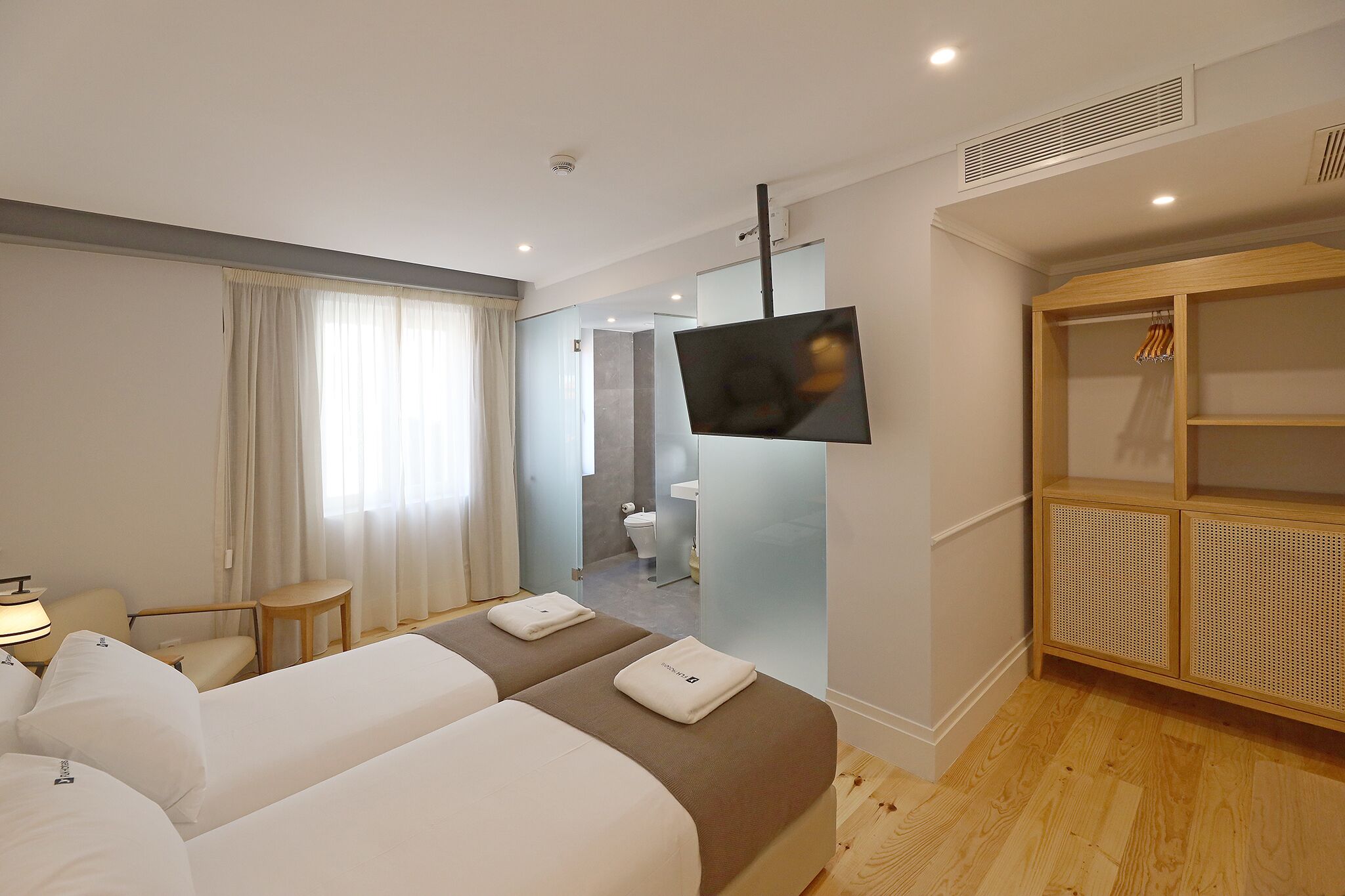 Habitación de hotel con dos camas, televisor, armario y baño visible a través de una pared de vidrio.