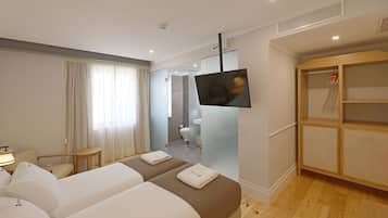 Habitación de hotel con dos camas, televisor, armario y baño visible a través de una pared de vidrio.