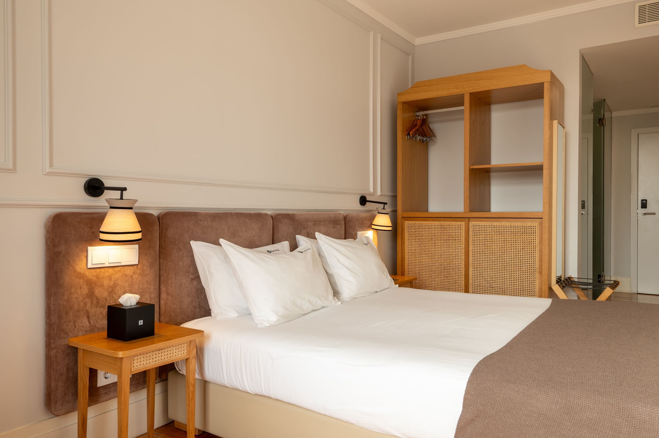 Una camera d'albergo con un letto grande, due lampade ai comodini, una testiera in legno, un tavolino con una scatola di fazzoletti e un armadio in legno con uno specchio.